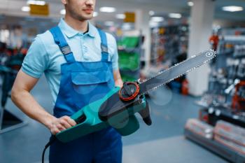 Male worker in uniform holds electric saw in tool store. Choice of professional equipment in hardware shop, instrument supermarket
