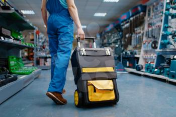 Male worker in uniform holds toolbox on wheels in tool store. Choice of professional equipment in hardware shop, instrument supermarket