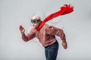 Elderly man in a red scarf and pilot glasses flying like a superman, grey background. Cheerful mature senior looking at camera in studio