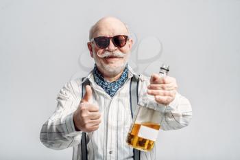 Fashionable elderly man holds the bottle of good alcohol, grey background. Mature senior looking at camera in studio, dude