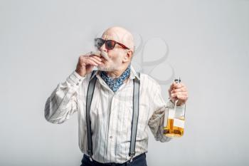 Fashionable elderly man with cigar and bottle of good alcohol, grey background. Mature senior looking at camera in studio, dude