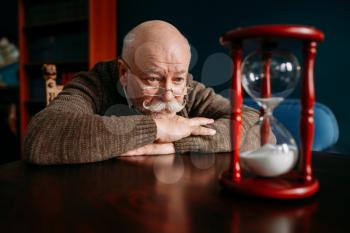 Serious elderly man looking on sandglass in home office, time cannot be turned back. Mature senior looking on hourglass
