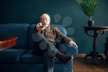 Elderly man watching TV on couch in home office. Bearded mature senior poses in living room, old age businessman