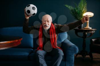 Elderly man with red scarf and ball watching TV, football fan. Bearded mature senior poses in living room, old age people leisure