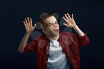 Little boy makes face leaning against the glass in studio. Happy childhood, children having fun, funny kid isolated on dark background, child emotion