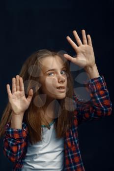 Little girl makes face leaning against the glass in studio. Happy childhood, children having fun, funny kid isolated on dark background, child emotion