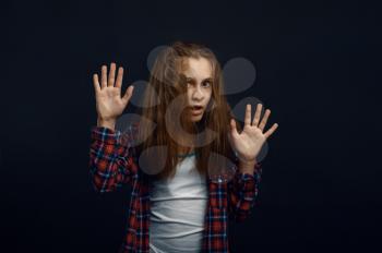Little girl makes face leaning against the glass in studio. Happy childhood, children having fun, funny kid isolated on dark background