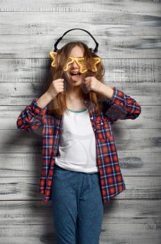 Funny little girl in headphones and star glasses in studio. Children and gadget, kid isolated on wooden background, child emotion, schoolgirl photo session
