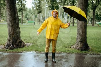 Man with umbrella walking in summer park in windy rainy day. Male person in rain cape and rubber boots, wet weather in alley