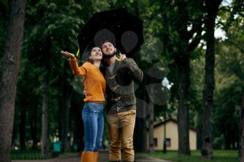 Happy love couple enjoys summer rainy day. Man and woman stand under umbrella in rain, romantic date on walking path, wet weather in alley
