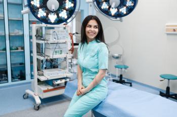 Smiling female surgeon poses at the operating table, lamp on background, surgery. Doctor in uniform, medical clinic worker, medicine and health, professional healthcare in hospital