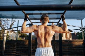 Muscular man does pull-ups exercise on a horizontal bar, street workout. Fitness training on sports ground outdoor, male person pumps muscles, active urban lifestyle