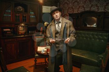 Male detective in hat and coat holds handcuffs at the crime scene, retro style. Criminal investigation, inspector with bracelets, vintage room interior on background