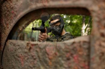 Paintball player in uniform and mask playing on playground in the forest. Extreme sport with pneumatic weapon and paint bullets or markers, military team game outdoors, combat tactics