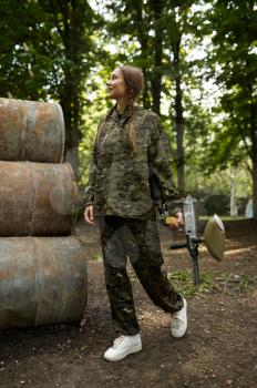 Female warrior with paintball gun poses on rusty barrels in the forest. Extreme sport with pneumatic weapon and paint bullets or markers, military team game outdoors, combat tactics