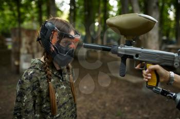 A shot from a paintball gun in the girl's face in mask, playground in the forest on background. Extreme sport with pneumatic weapon and paint bullets or markers, military team game outdoors