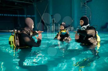 Instructor and two divers in suits, lesson in diving school. Teaching people to swim underwater with scuba gear, indoor swimming pool interior on background, group training