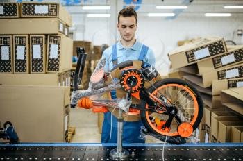 Bicycle factory, worker packs teen bike. Male mechanic in uniform installs cycle parts, assembly line in workshop, industrial manufacturing