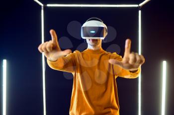 Young gamer plays the game using virtual reality headset and gamepad in luminous cube, front view. Dark playing club interior, spotlight on background, VR technology with 3D vision