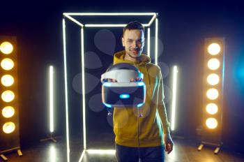 Young gamer poses with virtual reality headset and gamepad in luminous cube, front view. Dark playing club interior, spotlight on background, VR technology with 3D vision