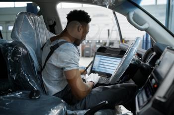 Male mechanic using laptop at the auto with opened hood, car service. Vehicle repairing garage, man in uniform, automobile station interior on background
