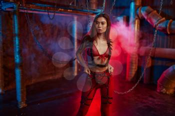 Sexy woman in red bdsm suit chained up, abandoned factory interior on background. Young girl in erotic underwear, sex fetish, sexual fantasy