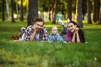 Mother, father and little baby leisures on grass in summer park. Mom and dad with male kid on lawn, picnic with child on plaid in the forest, family happiness