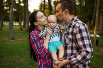 Mother and father kissing their little baby in summer park. Mom and dad with male kid on lawn, parents on picnic with child in the forest, family happiness