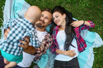 Mother, father and little baby lying on grass, top view. Mom and dad with male kid on lawn, parents on picnic with child in the forest, family happiness
