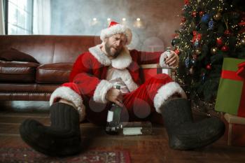 Vile drunk Santa claus sleeps under christmas tree with bottle of alcohol, nasty party, humor. Unhealthy lifestyle, bearded man in holiday costume, new year and alcoholism