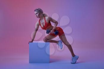 Sexy female athlete in red sportswear poses with dumbbell at the cube in studio, neon background. Fitness sportswoman at the photo shoot, sport concept, active lifestyle motivation