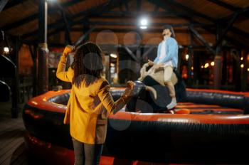 Love couple having fun on rodeo attraction in night amusement park. Family relax on fairground outdoors. Man and woman leisures on carousel, entertainment theme