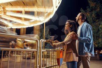 Love couple at the carousel in motion, night amusement park. Man and woman relax outdoors, roundabout attraction with lights on background. Family leisures in summertime, entertainment theme