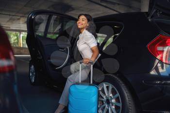 Happy woman with suitcase gets into the car on parking. Female traveler with luggage in vehicle park lot, passenger with bag. Girl with baggage near automobile