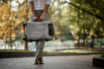 Woman poses with suitcase on walkway in summer park. Female traveler with luggage leisures outdoors, passenger with bag resting in nature. Girl with baggage relax on city alley