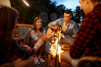 Friends playing on guitar by the bonfire, picnic at camping in the forest. Youth having summer adventure on rv, camping-car on background. Two couples leisures