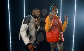 Two stylish rappers poses in glowing cube, studio with dark background. Hip-hop performers, break-dancers