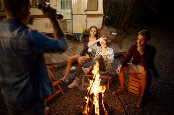 Man tells a scary story to his friends by the bonfire, picnic at camping in the forest. Youth having summer adventure on rv, camping-car on background. Two couples leisures, travelling with trailer