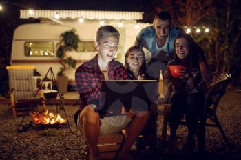 Friends with laptop by the campfire in the night, picnic at camping in the forest. Youth having summer adventure on rv, camping-car on background. Two couples leisures, travelling with trailer