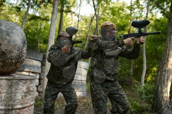 Paintball players in uniform and masks playing on playground in the forest. Extreme sport with pneumatic weapon and paint bullets or markers, military team game outdoors, combat tactics
