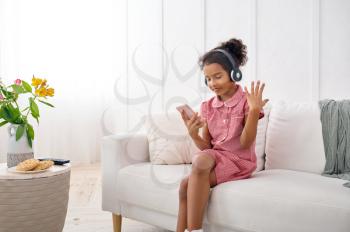 Little girl in headphones listening to music at home. Female kid sitting on sofa and using mobile phone, funny child with modern gadget