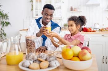Happy father and little kid drinks fresh juice on breakfast. Smiling family eats on the kitchen in the morning. Dad feeds female child, good relationship