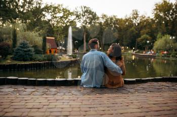 Love couple embracing at the pond in summer park. Man and woman relax outdoors, green lawn on background. Family hugging near the lake in summertime, weekend in nature