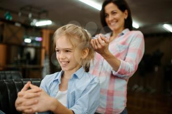Mother doing hairstyle to her daughter in salon. Mom and little girl play stylists together, happy childhood, glamour family