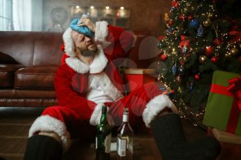 Vile Santa claus with hangover sitting under christmas tree with bottle of alcohol, nasty party, humor. Unhealthy lifestyle, bearded man in holiday costume, new year and alcoholism