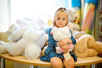 Pretty little girl poses with soft toys in kid's shop. Adorable child sitting on showcase in toyshop, happy childhood, baby makes a purchase in store
