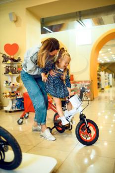 Mom and pretty little girl buying bicycle in kid's store. Mother and adorable daughter near the showcase in toyshop, happy childhood, family makes a purchase in shop