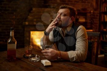 Man smokes cigarette and drinks alcohol beverage, bookshelf and rich office interior on background. Tobacco smoking culture, specific flavor