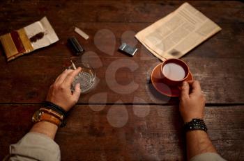 Man with cup of coffee smokes a cigarette at wooden table, top view. Tobacco smoking culture, specific flavor. Male smoker leisures