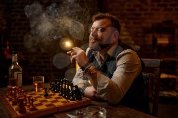 Man smokes a cigar, drinks alcohol beverage and play chess, bookshelf and vintage office interior on background. Tobacco smoking culture, specific flavor. Male smoker leisures at the chessboard